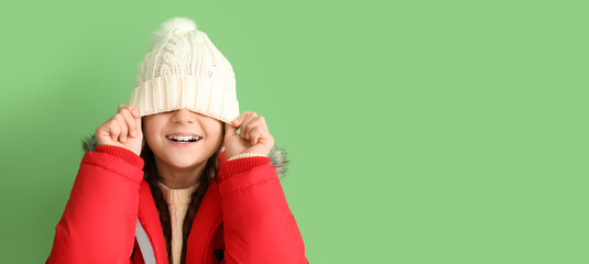Wall Mural - Funny little girl in winter hat and warm coat on green background with space for text
