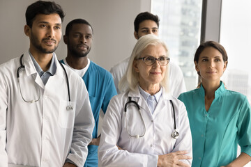 Wall Mural - Multiethnic different aged group of doctors standing together for shooting, looking away. Confident senior older head of clinic posing in front of team with hands folded