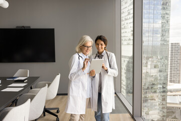 Wall Mural - Two medical professional women discussing online medical service on tablet together, using digital device, talking, Happy younger and older doctors meeting in boardroom