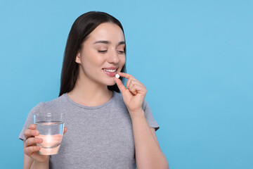 Poster - Beautiful young woman with glass of water taking pill on light blue background, space for text