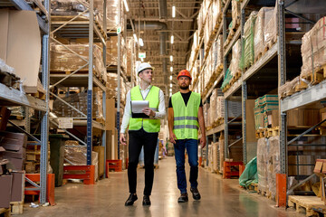 Retail Warehouse full of Shelves with Goods, Two Young Caucasian Male Workers Supervisors in Working Uniform and Helmet Discuss Product Delivery. Men Walking and Having Talk In Warehouse