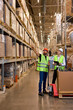 Caucasian warehouse managers walking through large warehouse distribution center discussing about increasing productivity. good teamwork, dressed in working clothes vest and hard hats