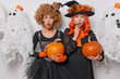 © wayhome.studio  - Horizontal shot of shocked two women stare with scared expressions wear black dress hold ripe orange pumpkins going to carve them notice something spooky surrounded by ghosts against white background