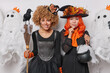 © wayhome.studio  - October enchantment. Two women stand closely to each other with mysterious expressions hold broom cauldron pose at halloween party prepare for upcoming October 31st carnival make spooky decorations