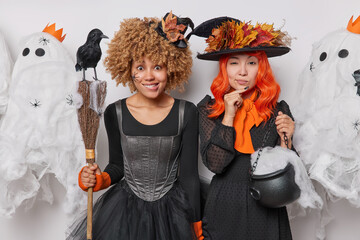 Wall Mural - October enchantment. Two women stand closely to each other with mysterious expressions hold broom cauldron pose at halloween party prepare for upcoming October 31st carnival make spooky decorations