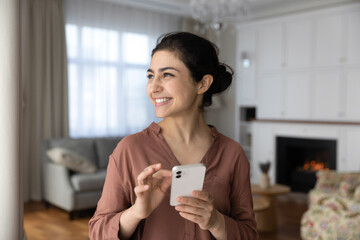 Wall Mural - Dreamy smiling Indian woman holding smartphone, looking in distance, standing at home, happy pensive young female distracted from online chatting, visualizing good future, planning and dreaming