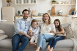 © fizkes - Cheerful young attractive couple with little children sit on couch in studio apartment smile, pose, look at camera, hug, show love, unity and affection. Happy homeowners family portrait, parenthood