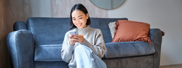 People and technology. Smiling asian woman sitting at home, using her mobile phone, typing message, browing internet or shopping online from smartphone app