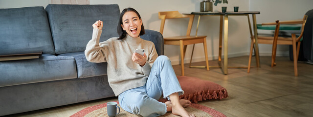 Wall Mural - Enthusiastic asian woman sits on floor with smartphone, raise hand up and cheering, celebrating victory