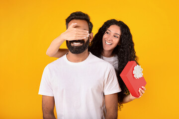 Lady closes eyes to husband surprising giving gift, yellow background