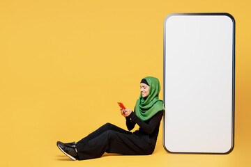 Wall Mural - Full body young asian muslim woman wears green hijab abaya black clothes big huge blank screen mobile cell phone use smartphone isolated on plain yellow background. People uae islam religious concept.