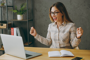 Wall Mural - Young overjoyed excited happy successful employee business woman wears shirt casual clothes glasses sit work at office desk with pc laptop computer do winner gesture. Achievement career job concept.