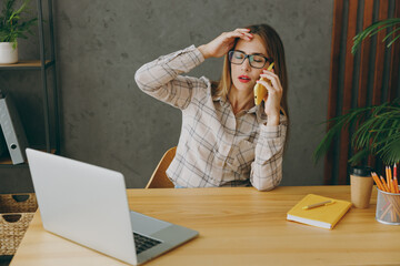 Wall Mural - Young exhausted sad employee business woman wearing shirt casual clothes glasses sit work at office desk with pc laptop computer talking speaking on mobile cell phone. Achievement career job concept.