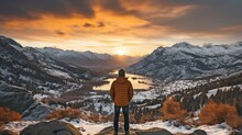 Person Standing On A Snow-covered Ledge Overlooking The Stunning Sunset Vista Of Mountains.