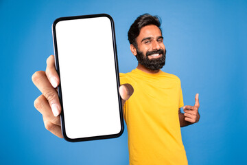 Poster - Online ad. Smiling middle aged indian man pointing at blank smartphone in his hand, blue background, mockup