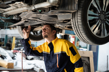 Vehicle mechanic conduct car inspection from beneath lifted vehicle. Automotive service technician in uniform carefully diagnosing and checking car's axles and undercarriage components. Oxus