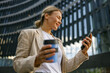 © Yaroslav Astakhov - Smiling woman office worker is standing on modern building background and looking on phone