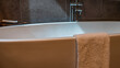 Details of the bathroom interior. A terry towel is draped over the edge of a large white acrylic bathtub. Close-up. Metal taps on the beige tiled wall.