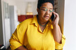 © shurkin_son - Serious african american senior woman in glasses and yellow shirt talking on phone with serious worried facial expression, receiving bad news from her doctor or relatives, leaning on wall