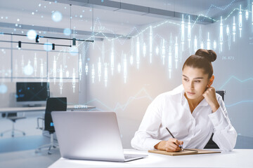 Canvas Print - Attractive young european businesswoman sitting at desk and using laptop with creative glowing forex chart hologram on blurry office interior background. Trade and finance concept.