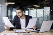 © Tetiana - A young man office worker sits in the office at the table, holds papers in his hands, looks confusedly at documents and bills