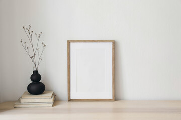 Black organic shaped vase with dry flowers, grass on old books. Blank vertical wooden picture frame mockup. Home office table. Minimal Scandi boho interior. White wall background. Empty copyspace.