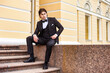 © Alex Vog - Serious handsome guy in tuxedo sitting on stairs, strong looking at camera. Confident businessman young man in tux posing at urban street outdoors, luxury image cinematic style. Copy ad text space