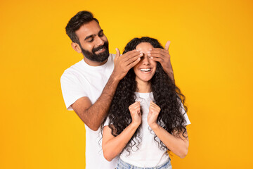 Wall Mural - Middle eastern man covering wife's eyes surprising her, yellow background