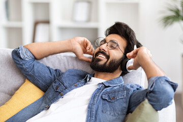 Wall Mural - Young Smiling Indian Guy Wearing Wireless Headphones Listening Music At Home