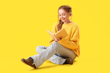 Poster - Beautiful happy young woman in warm sweater sitting on yellow background and reading book