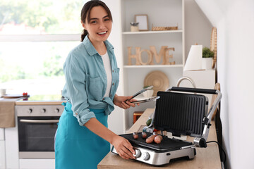 Canvas Print - Beautiful young woman cooking delicious sausages on modern electric grill in kitchen