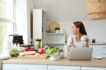 Poster - Beautiful young woman using laptop while cooking delicious sausages on modern electric grill in kitchen