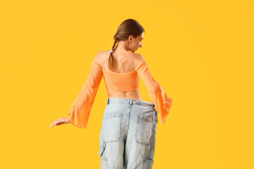 Canvas Print - Beautiful young woman in loose jeans on yellow background, back view. Weight loss concept