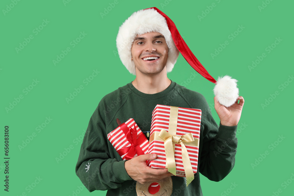 Happy young man in Santa hat with gift boxes on green background