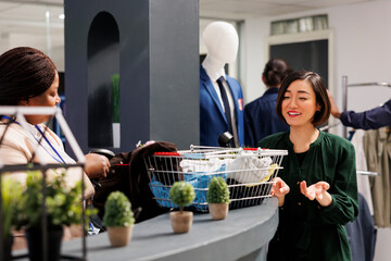Wall Mural - Happy satisfied Asian woman customer standing at checkout cashier counter in clothing store, buying new clothes in fashion outlet. Friendly retail sales assistant serving shopper