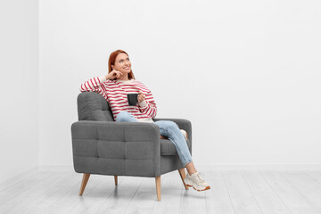 Poster - Happy young woman with cup of drink sitting in armchair near white wall indoors. Space for text