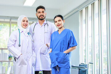 Asian Indian professional experienced bearded male doctor in white lab coat with stethoscope with female muslim colleague in hijab and surgeon in blue uniform standing smiling together in hospital