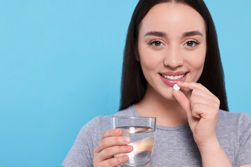 Canvas Print - Beautiful young woman with glass of water taking pill on light blue background, space for text