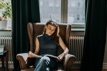 Teenage Girl Reading Book While Sitting On Armchair Against Window At Library