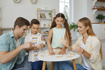 Family leisure. Cheerful young family with two children is having fun putting together wooden puzzles. Caucasian mom, dad, son and daughter play games together sitting in living room at small table.