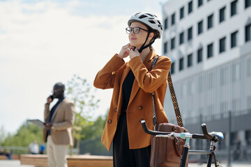 Poster - Happy young female employee fastening belt of protective cycling helmet while standing in front of camera by her bicycle in urban environment