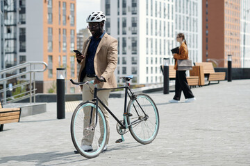 Wall Mural - Young businessman in safety helmet texting in smartphone in urban environment while standing by his bicycle against modern architecture