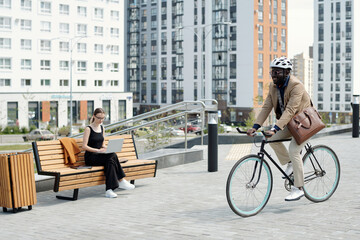 Wall Mural - Happy young businessman in formalwear and safety helmet moving by businesswoman with laptop while riding bicycle in urban environment