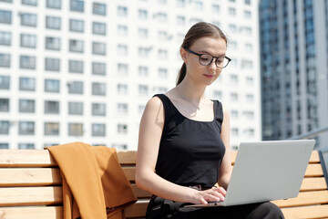 Wall Mural - Young businesswoman in eyeglasses and black vest typing and looking at laptop screen while sitting on bench and organizing work outdoors