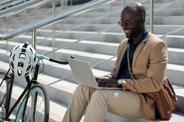 Young African American male solopreneur or freelancer typing on laptop keyboard while sitting on staircase and communicating in chat