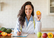 Attractive european nutrition adviser holding orange and making meal plan for client, working at weight loss clinic