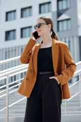 Poster - Young smiling businesswoman in brown blazer and black vest and pants talking to someone on smartphone while standing on staircase