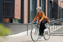 Young woman in formalwear and safety helmet sitting on bicycle and riding home from work while moving along modern building