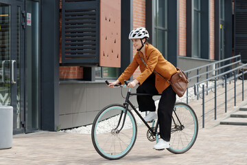 Poster - Side view of young businesswoman in quiet luxury attire and protective helmet riding bicycle along modern building of office center after work