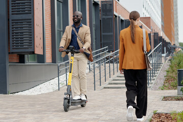 Poster - Rear view of young businesswoman in brown blazer and black pants passing by male employee in quiet luxury attire riding scooter